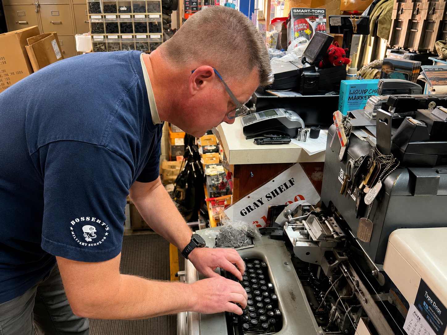 Don Bossert stamping a custom dog tag on the store's vintage embossing machine, finished tags hanging on ball chains beside it.