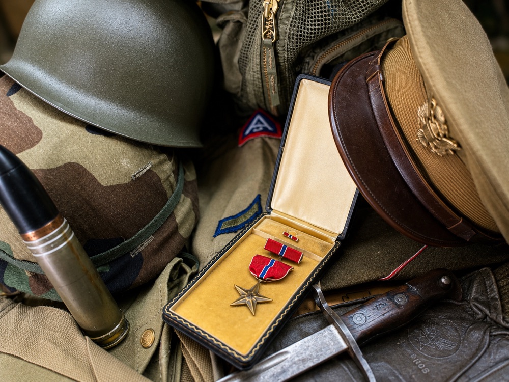 A spread of collectible militaria at Bossert's for identification and appraisal, an M1 helmet, a cased Bronze Star medal, a WWII officer's visor cap, unit patches, an artillery shell, and a bayonet.
